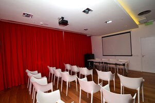 A conference room with a red curtain backdrop, white chairs, a projector screen, and a table with chairs.