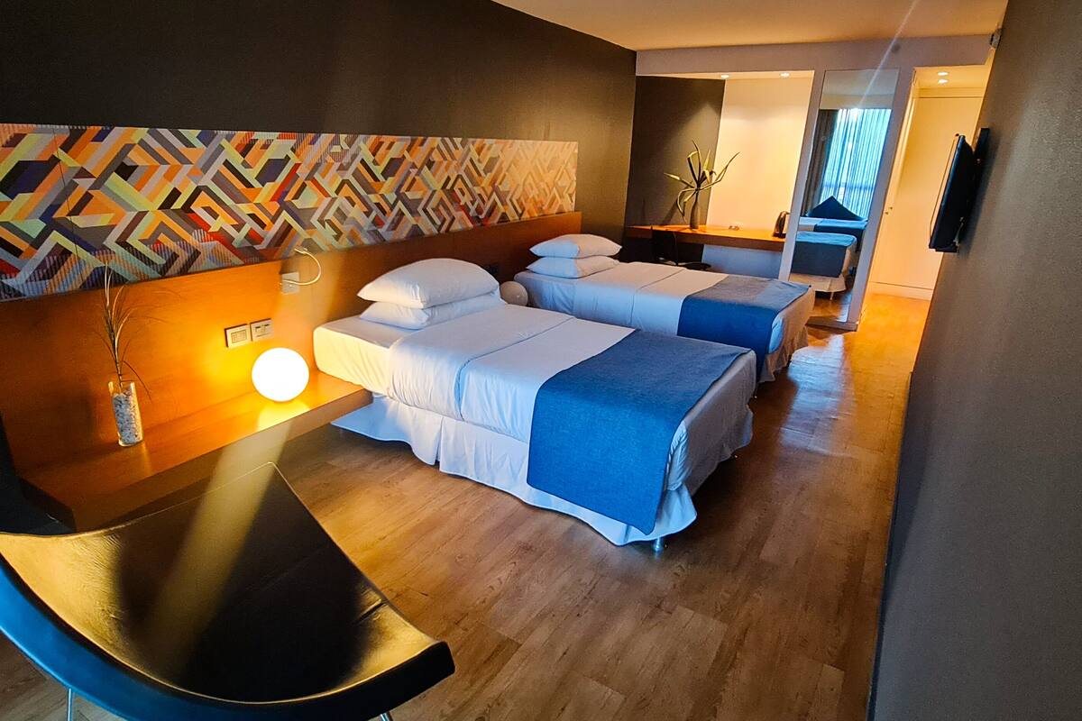 A hotel room with two beds, a desk, a chair, a wall with a geometric pattern, and a nightstand with a lamp.