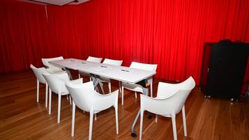 Une salle de conférence avec une table et des chaises blanches, des rideaux rouges et un chariot noir pour les haut-parleurs.
