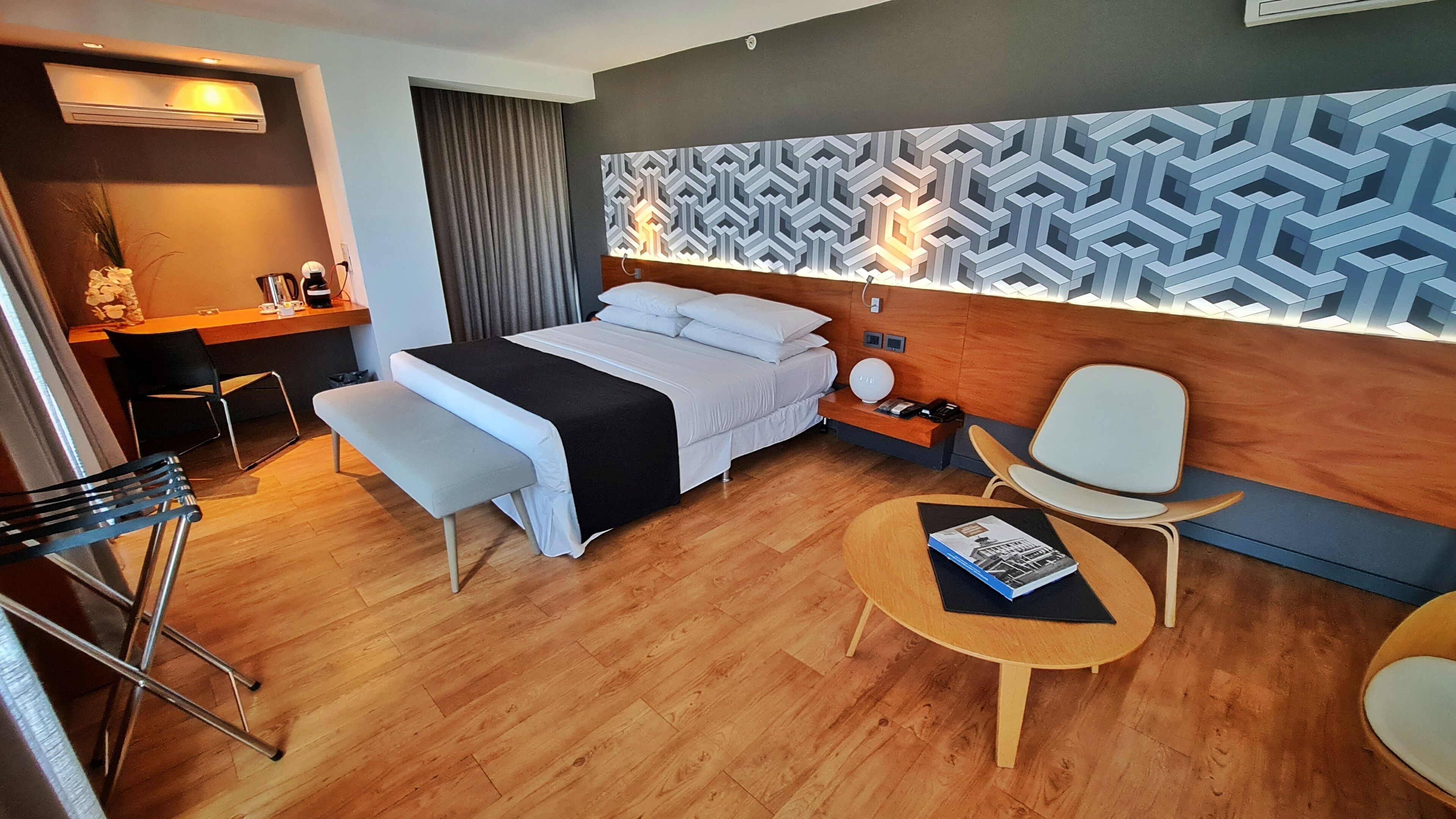 A modern hotel room with a wooden floor, a bed with white bedding, a desk with a chair, a small table with a book, and a patterned wall.