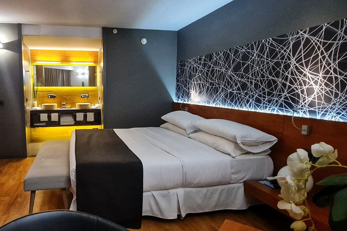 A modern hotel room with a bed, a chair, a bench, a vanity with two sinks, and a decorative wall.