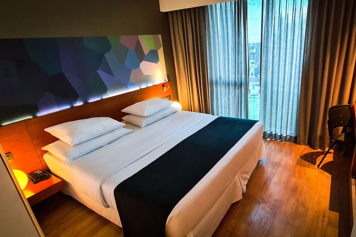 A hotel room with a large bed, a headboard with a geometric design, a bedside table with a lamp, and a view of the outdoors through curtains.