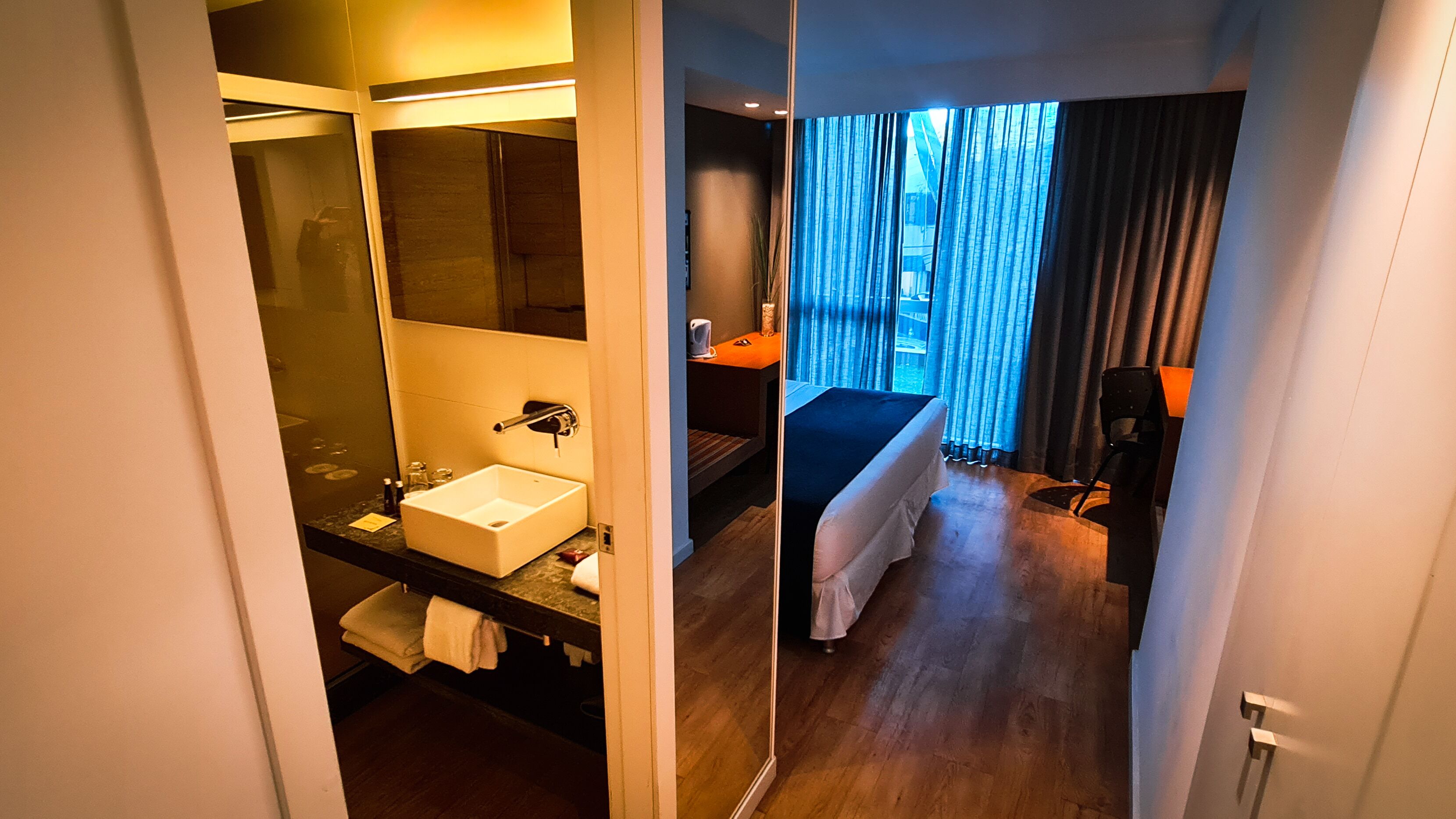 A hotel room with a large mirror, a sink, a bed, and a view of the city through the window.