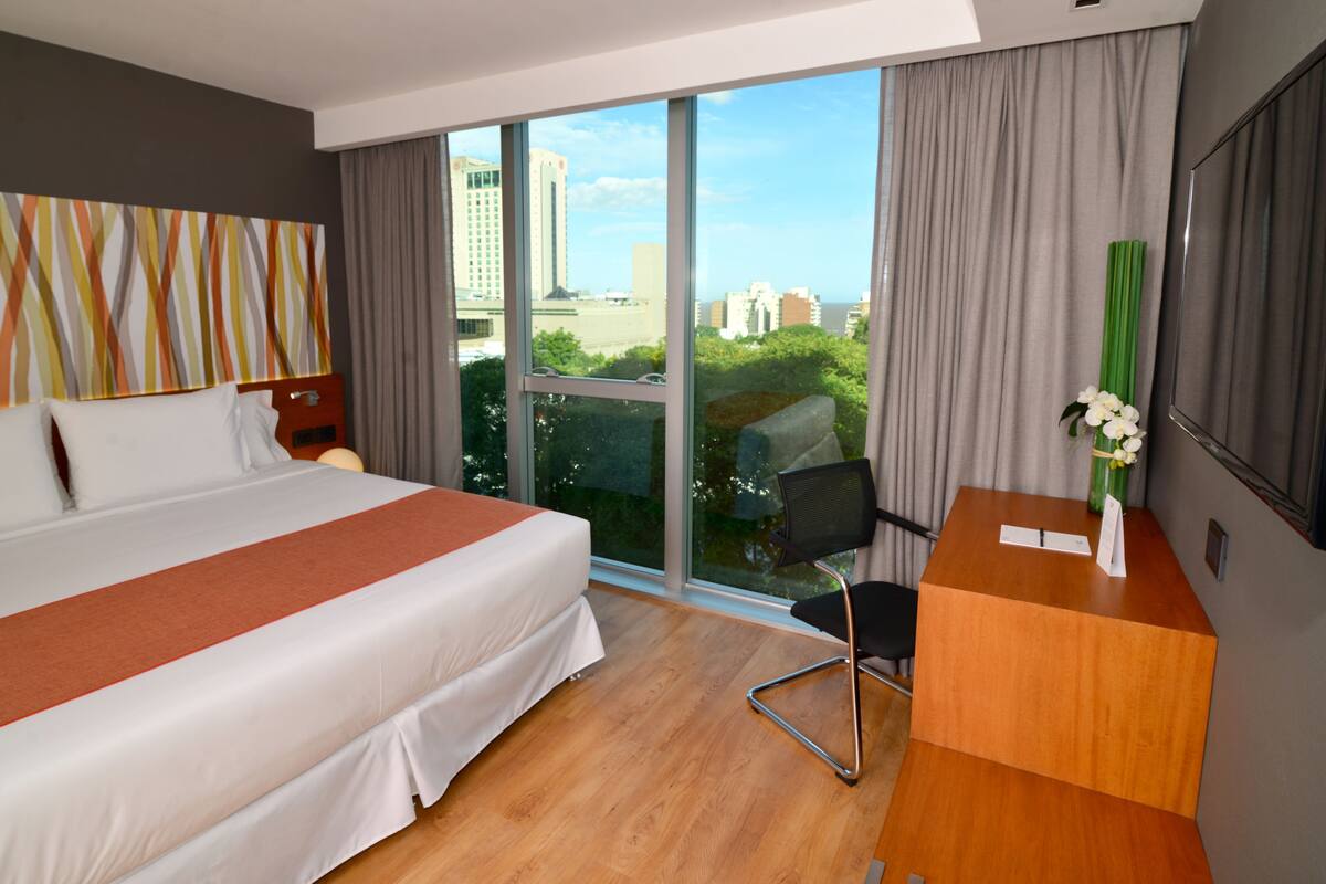 A modern hotel room with a large bed, a desk, and a view of the city.