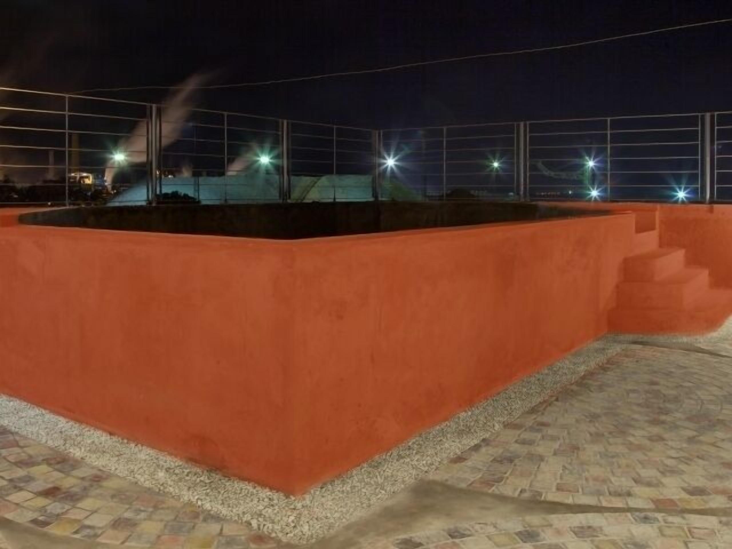 Un espacio exterior rectangular con piso de baldosas, rodeado por un muro bajo y una cerca metálica.