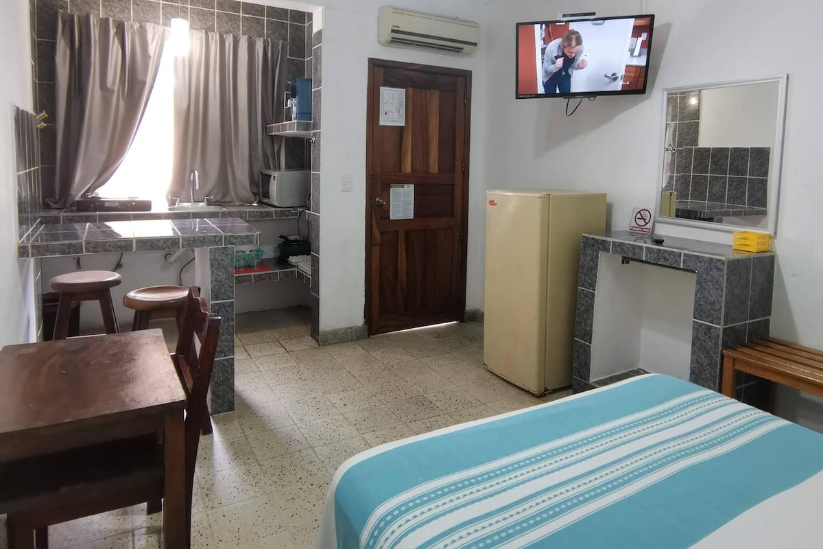Quarto compacto com cama, escrivaninha, cadeira, televisão e ventoinha no teto.