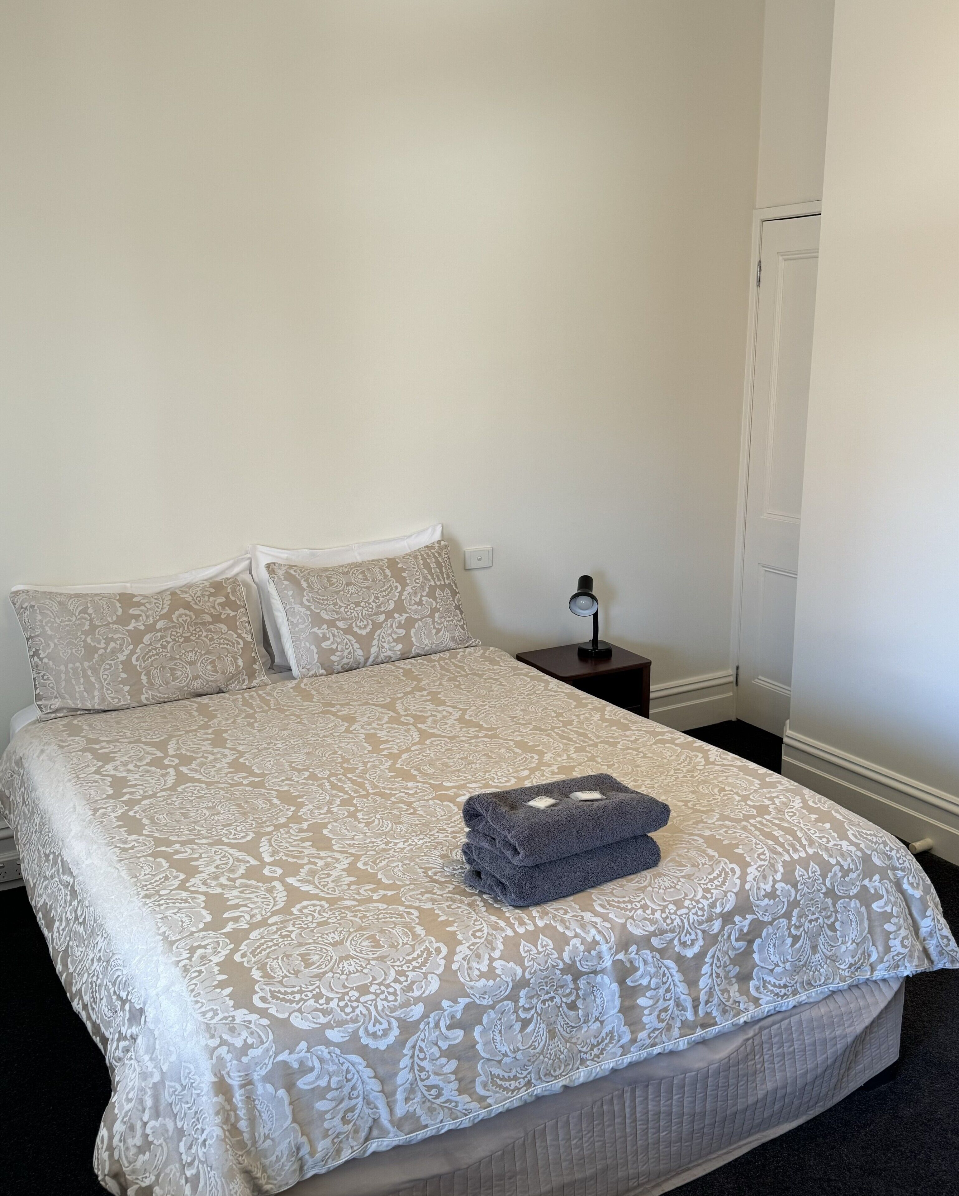 A neatly made bed with a patterned bedspread and pillows, a folded towel on the bed, and a nightstand with a lamp.