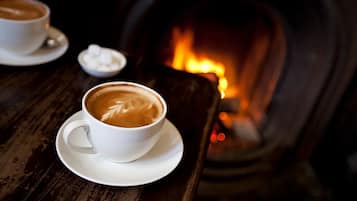 Une tasse de café ornée d’un motif en latte art, posée sur une soucoupe, sur une table en bois, avec une cheminée en arrière-plan.