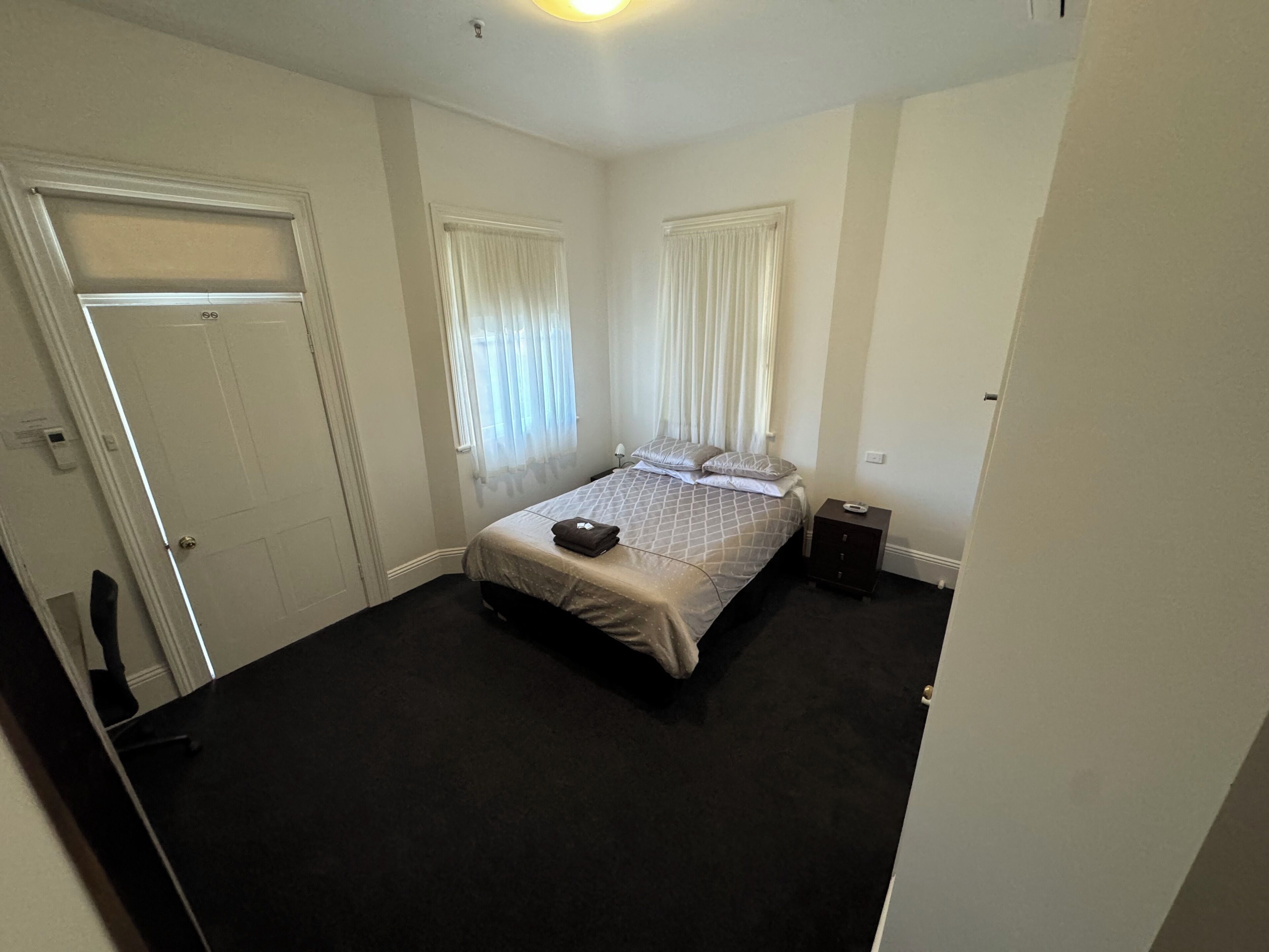 Un dormitorio con una cama, dos ventanas con cortinas, una mesita de noche y una puerta.