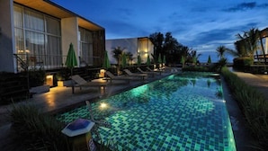 A swimming pool with a checkered pattern, surrounded by lounge chairs and umbrellas, and adjacent to a building with large windows and outdoor lighting. - LG Lemon Grass (Sattahip)