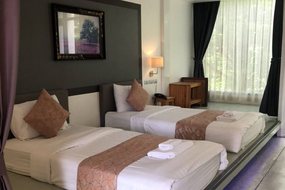 Une chambre d’hôtel avec deux lits, une table de chevet, une lampe, un tableau encadré au mur et une fenêtre avec des rideaux.