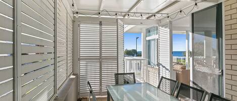 A balcony with a glass table and chairs, white shutters, and a view of the ocean.