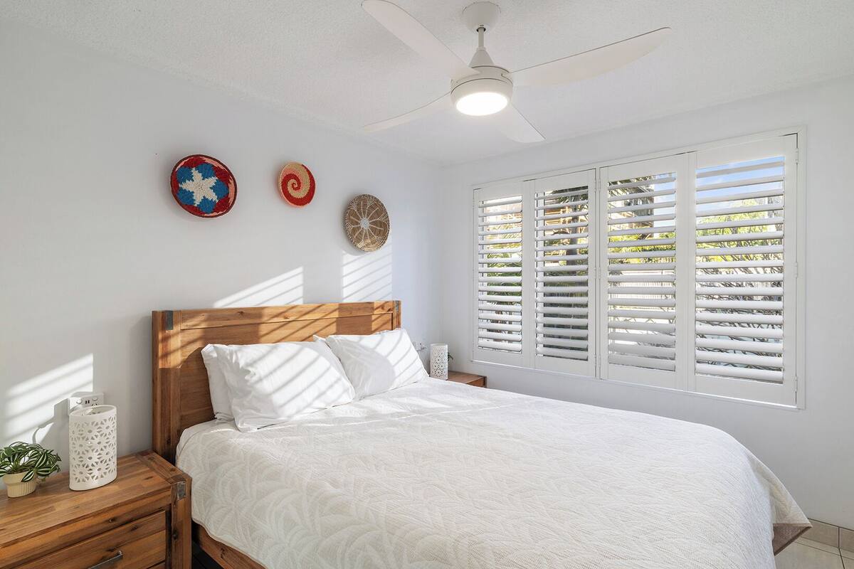 A bedroom with a wooden headboard, white bedding, a ceiling fan, and a window with shutters.