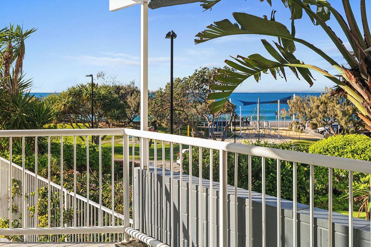 A balcony with a view of a beach, palm trees, and a blue building in the background.