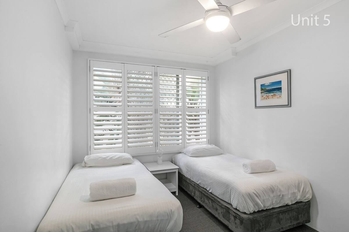 A bedroom with two single beds, a small bedside table, a ceiling fan, and a window with white shutters.