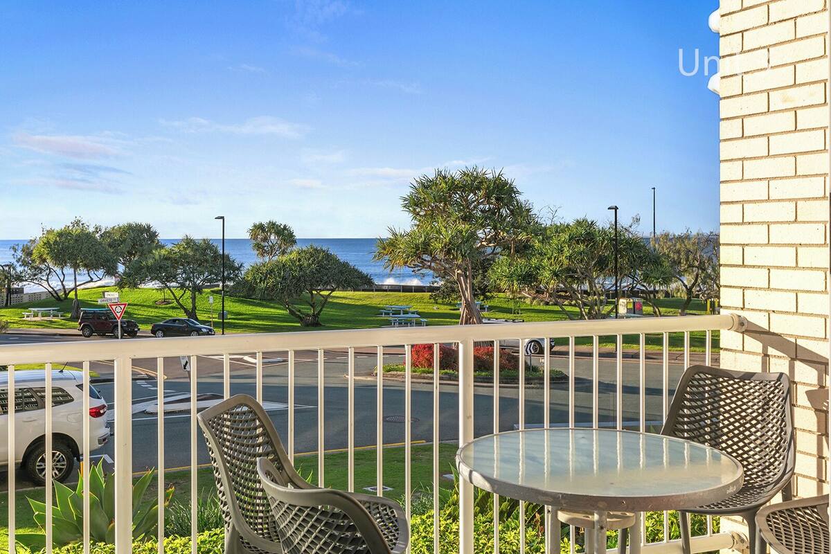 A balcony with two chairs and a table overlooking a road with cars and a grassy area with trees.