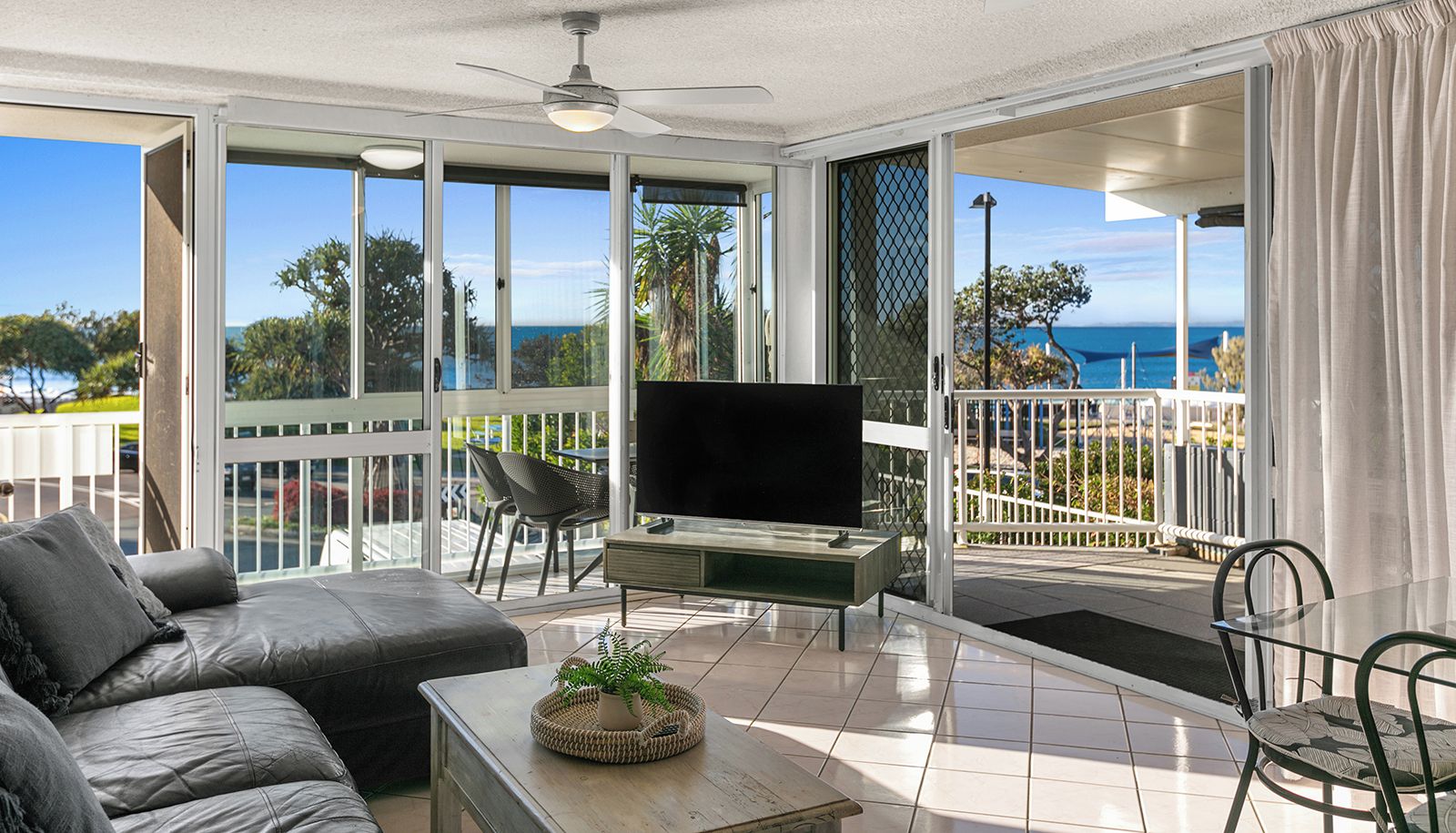 Ein modernes Wohnzimmer mit Flachbildfernseher, einem grauen Sofa und einem Esstisch mit Pflanze. Große Fenster bieten Blick auf das Meer und Palmen.