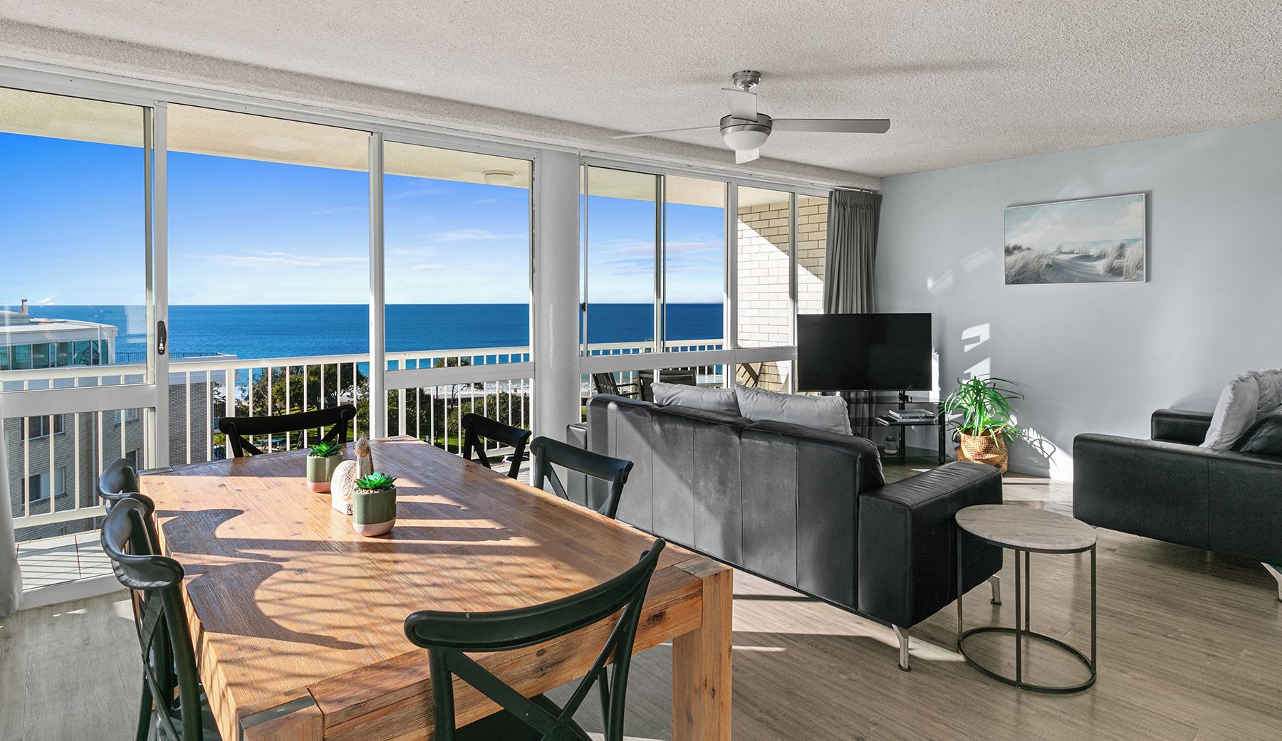 A spacious living area with a wooden dining table, black leather sofa, and a TV. Large windows offer a view of the ocean.