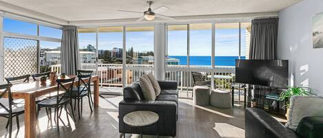 A spacious living room with a large window overlooking the sea, a ceiling fan, and a flat-screen TV.