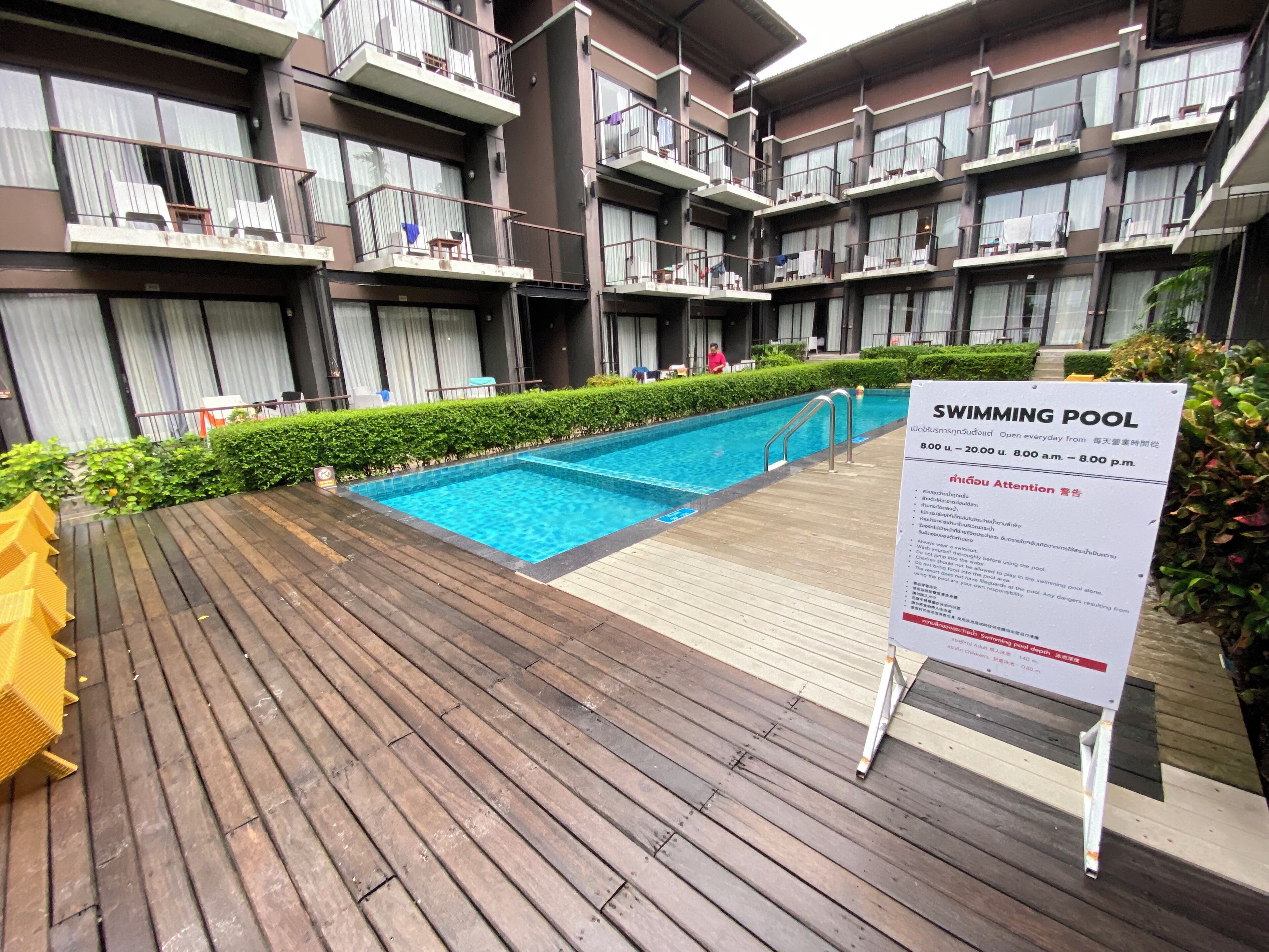 A swimming pool with a wooden deck, surrounded by a multi-story building with balconies.