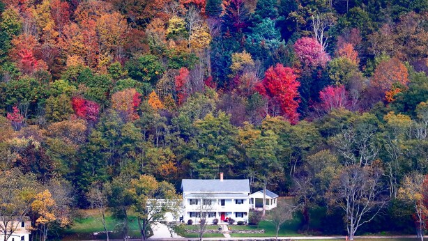 A white two-story house with a porch, surrounded by a forest with autumn-colored trees. - The Hann Homestead Inn (Andover)