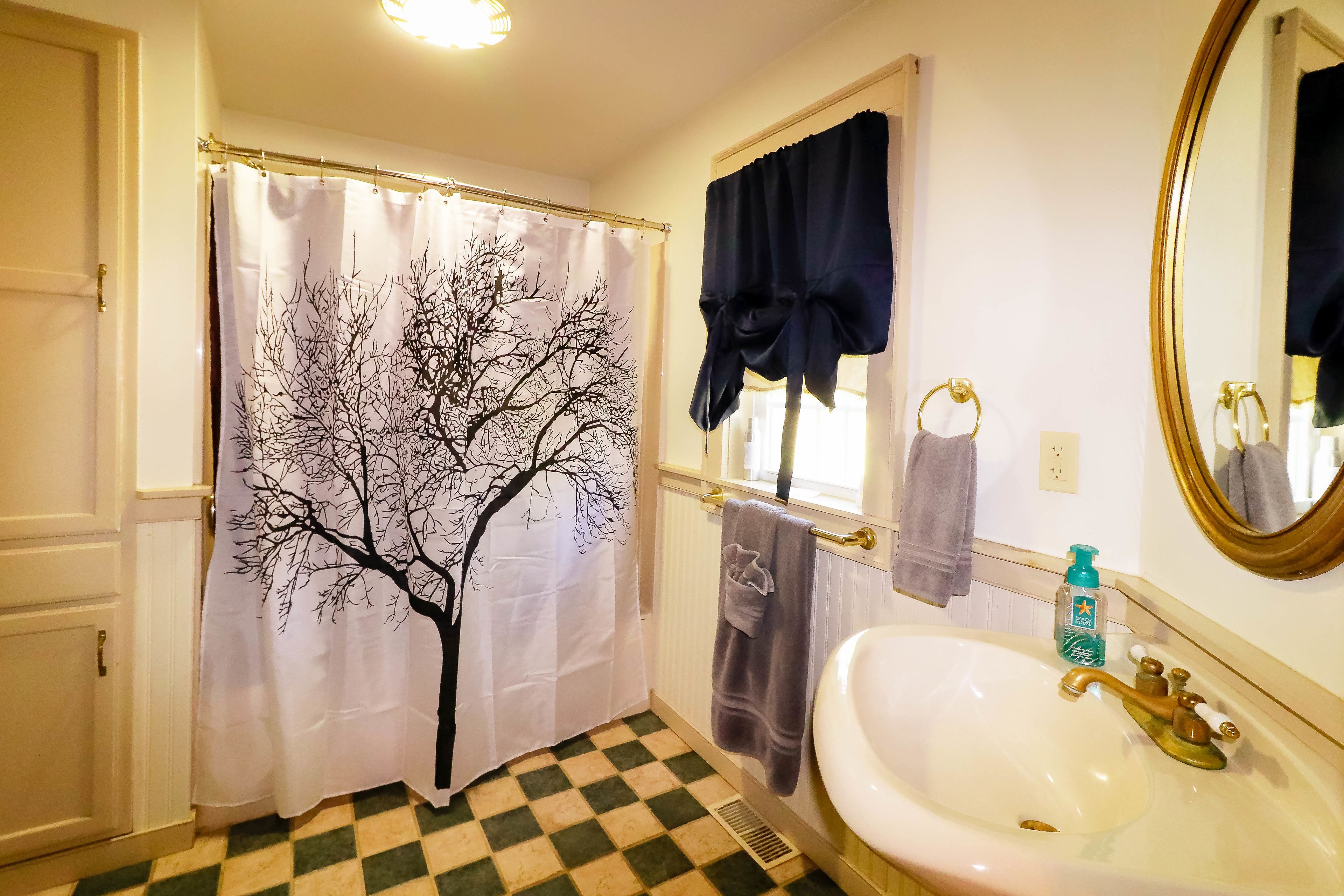 Un bagno con pavimento a scacchi, un lavandino bianco con rubinetteria dorata, uno specchio con cornice dorata, una finestra con tenda nera e una tenda da doccia con motivo a alberi.