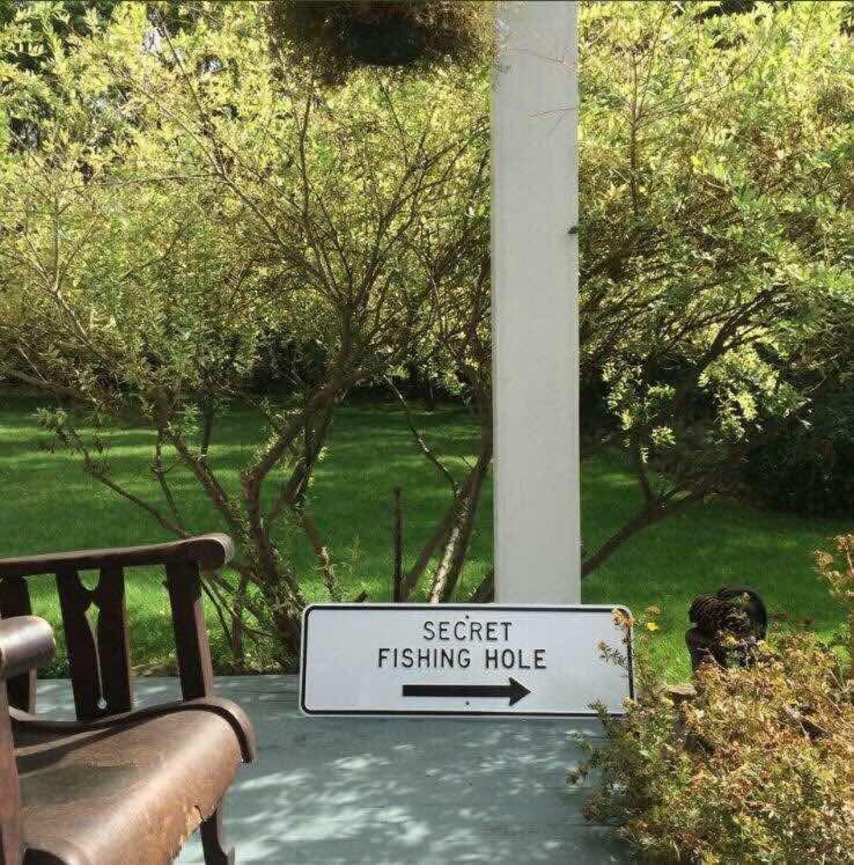 A sign for a 'Secret Fishing Hole' with an arrow pointing to the right, set against a backdrop of lush greenery and a bench.