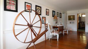 A room with a large wooden wheel, white chairs, a table, and various framed posters on the wall. - The Hann Homestead Inn (Andover)