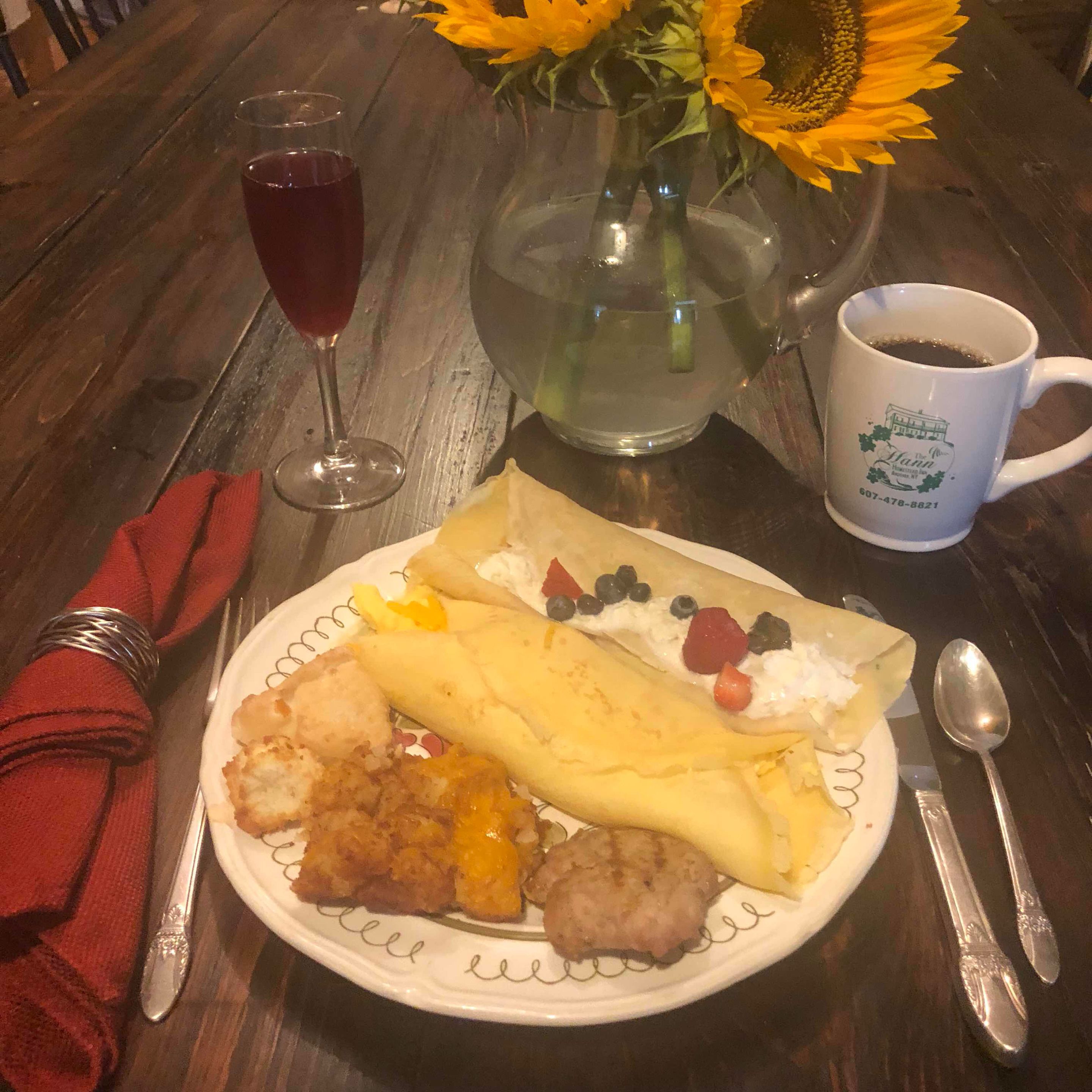 A plate with a crepe, meatballs, and a side dish, a glass of red drink, a coffee mug, and a vase with sunflowers.