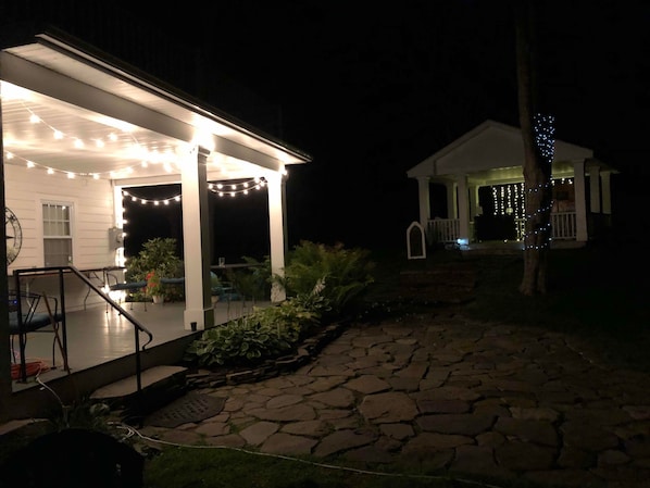 A covered outdoor patio area with string lights, a stone pathway, and a small white building with blue lights.