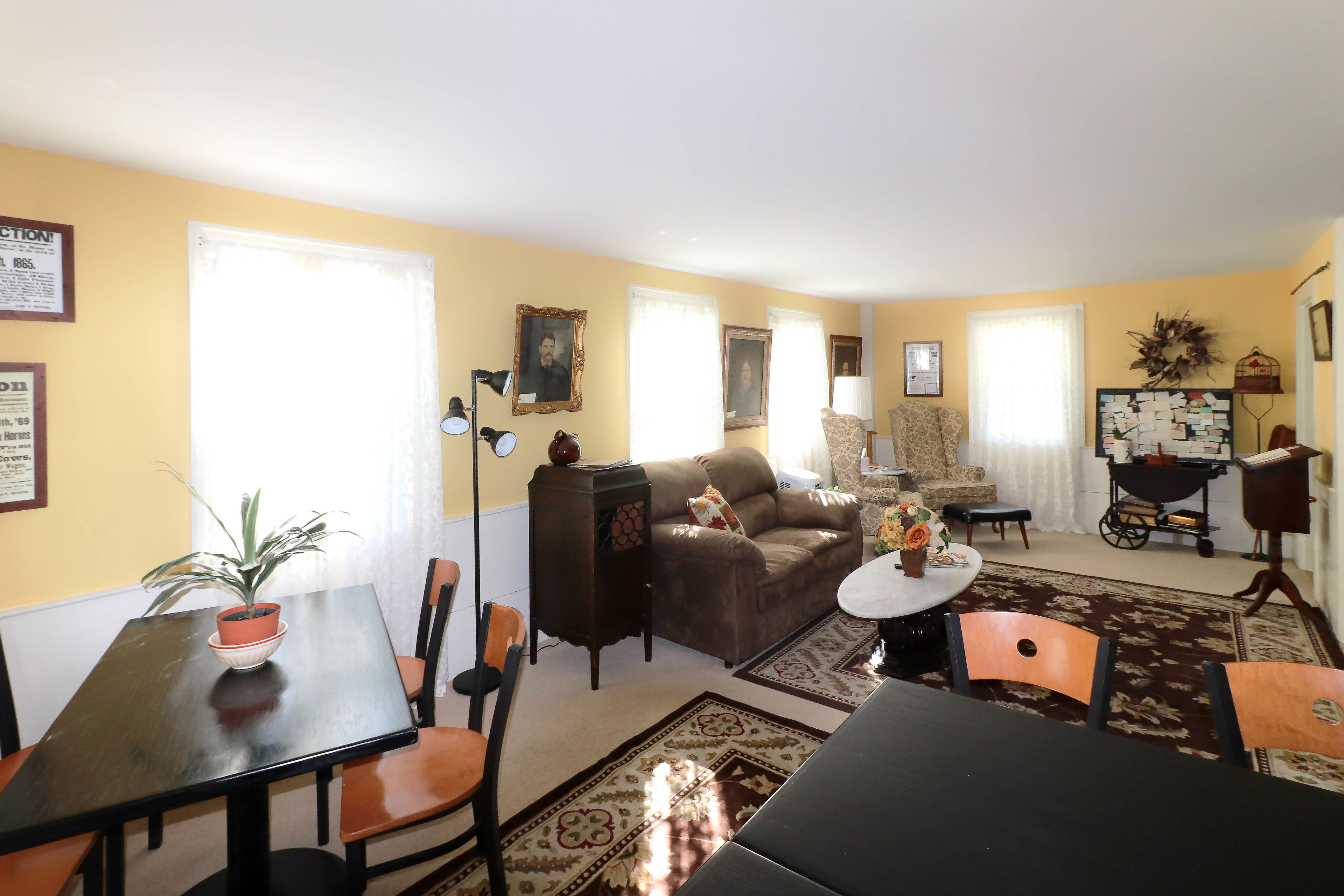 A living room with a brown sofa, a dining table with chairs, a potted plant, and a wall-mounted lamp.
