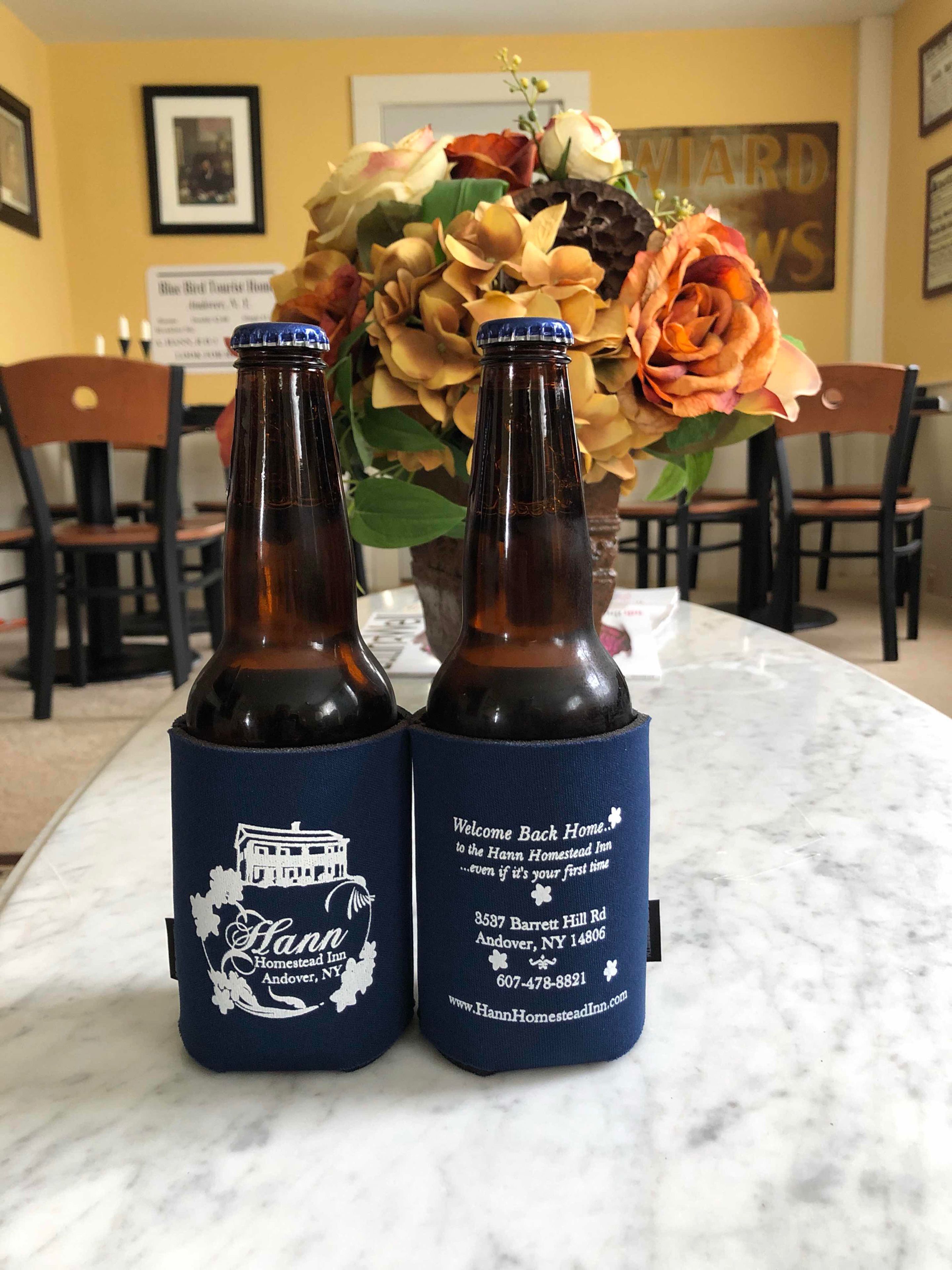 Two bottles with labels on a table, a vase with flowers, and a framed picture on the wall.