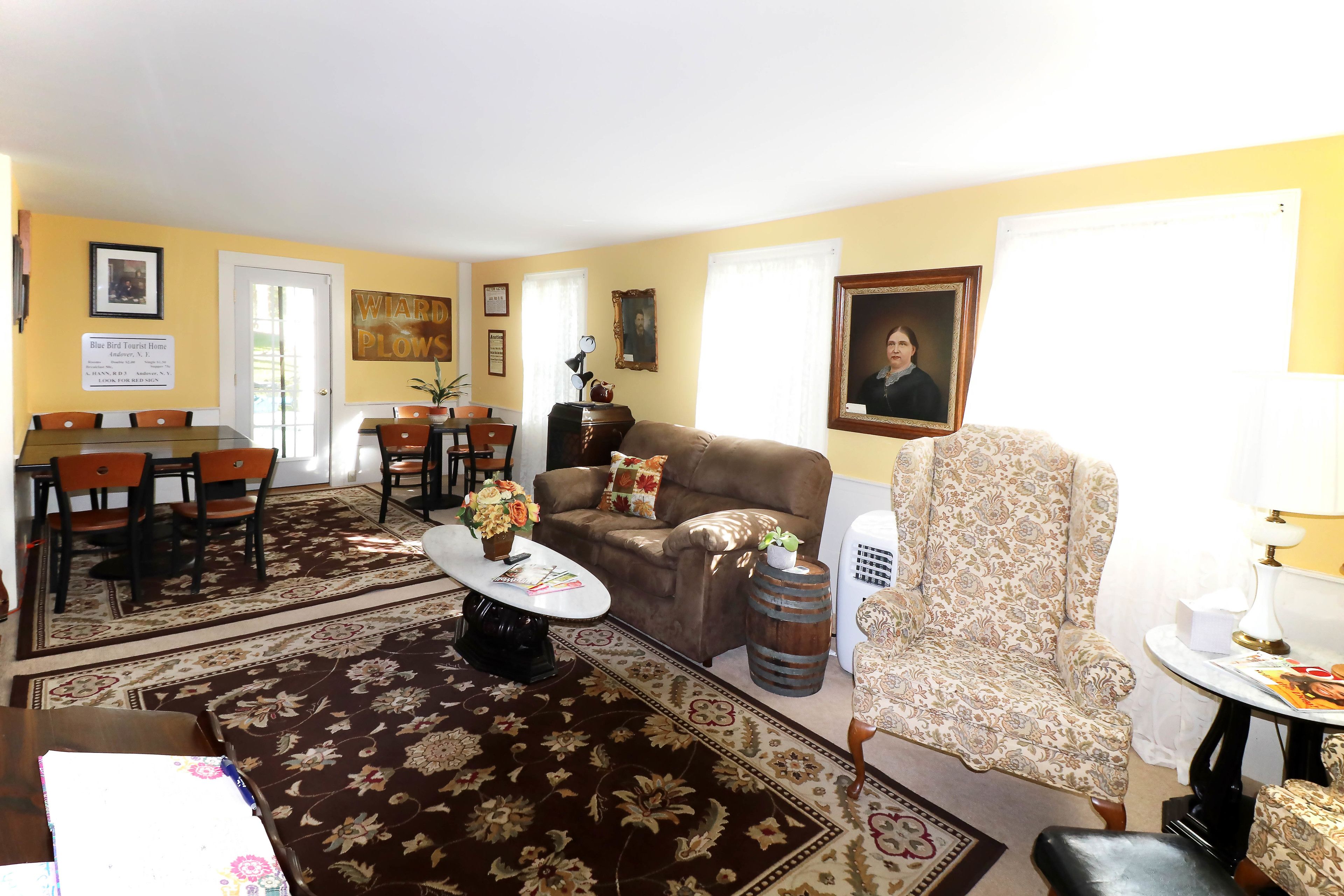 A living room with a brown sofa, a patterned armchair, a coffee table with a vase, a dining area with chairs, and a framed portrait on the wall.