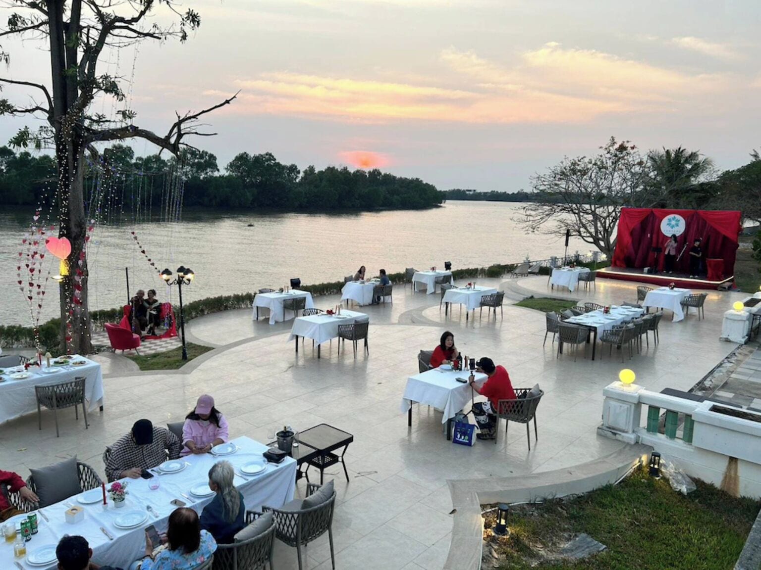 An outdoor dining area with tables and chairs arranged on a terrace overlooking a body of water, with a stage set up for an event.