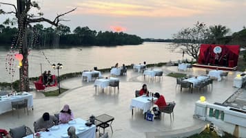 An outdoor dining area with tables and chairs arranged on a terrace overlooking a body of water, with a stage set up for an event.
