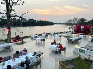 Un área de comedor al aire libre con mesas y sillas dispuestas en una terraza con vistas a un cuerpo de agua, y un escenario montado para un evento.