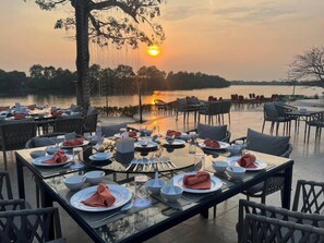 An outdoor dining area with tables set for a meal, overlooking a body of water and a sunset. - Suntara Wellness Resort & Hotel (Chachoengsao)