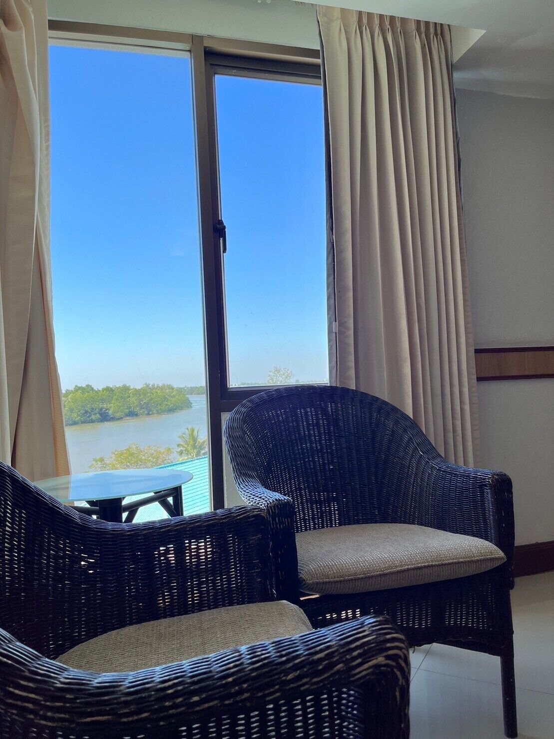 Ein Zimmer mit zwei Korbstühlen, einem Glastisch und Blick auf ein Gewässer durch ein großes Fenster mit Vorhängen.