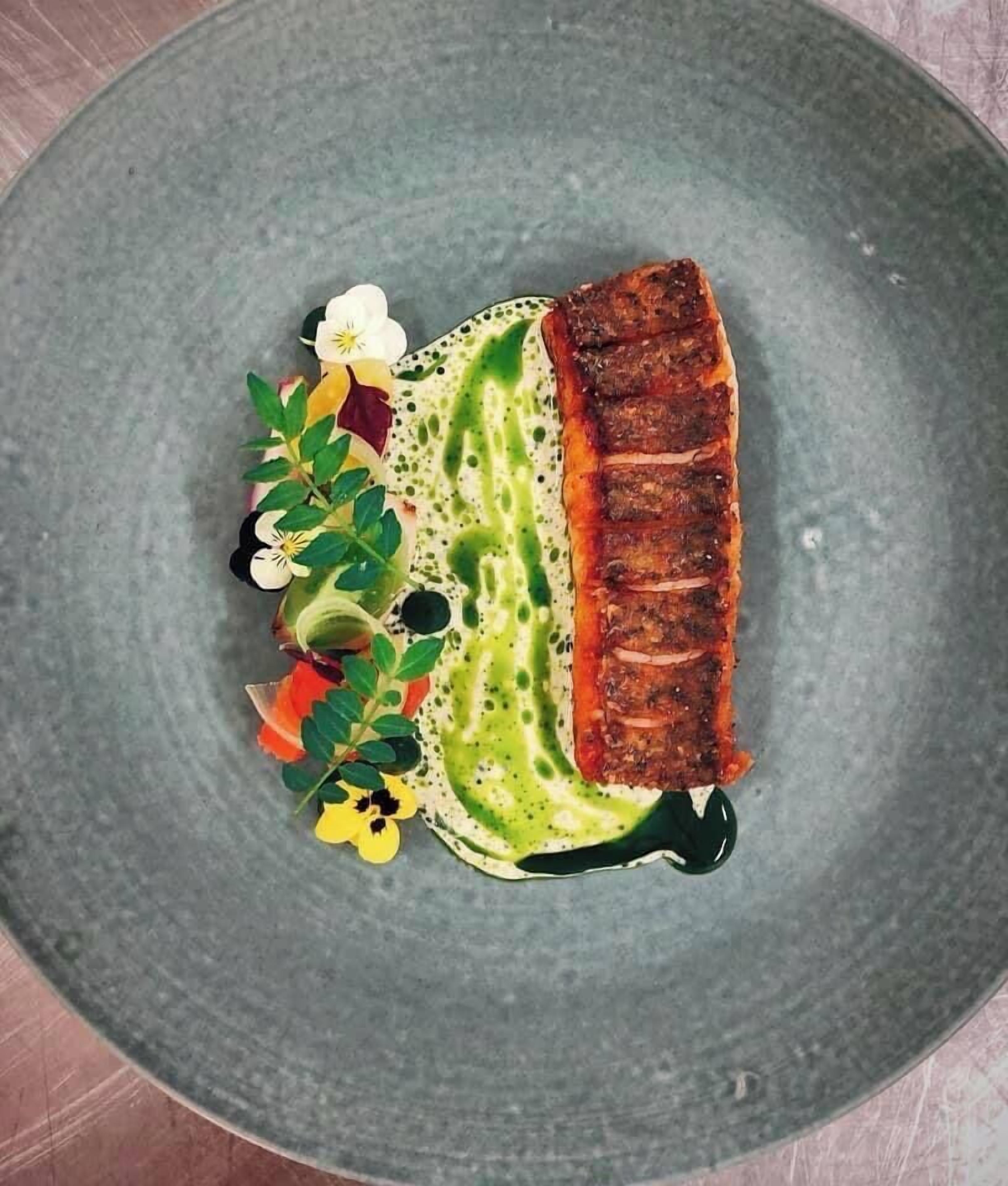 Un plato con un trozo de pescado a la parrilla, salsa verde y flores decorativas.