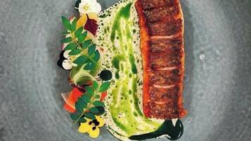 Un plato con un trozo de pescado a la parrilla, salsa verde y flores decorativas.