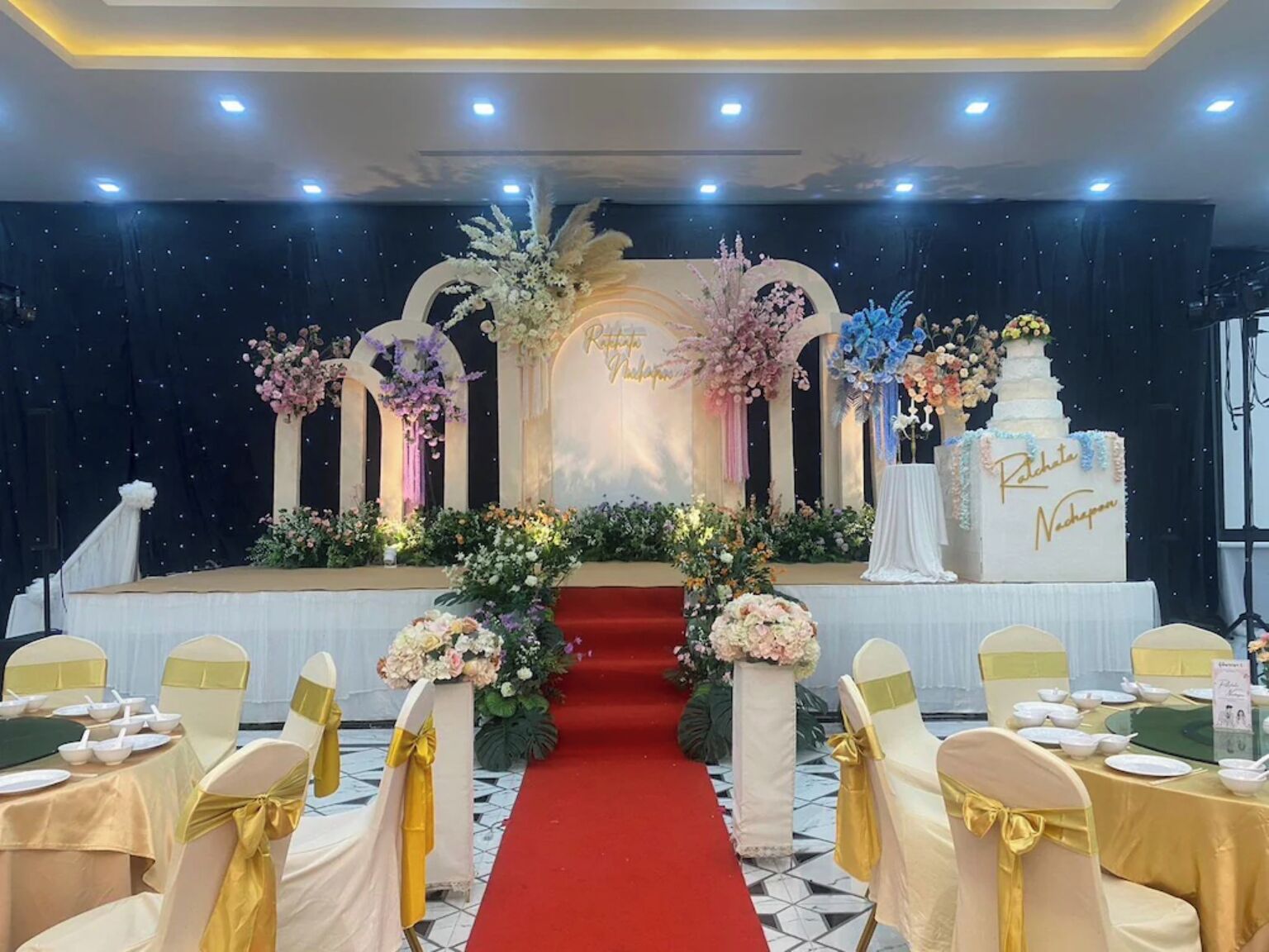 Un escenario con decoraciones florales, alfombra roja y un pastel con nombres.