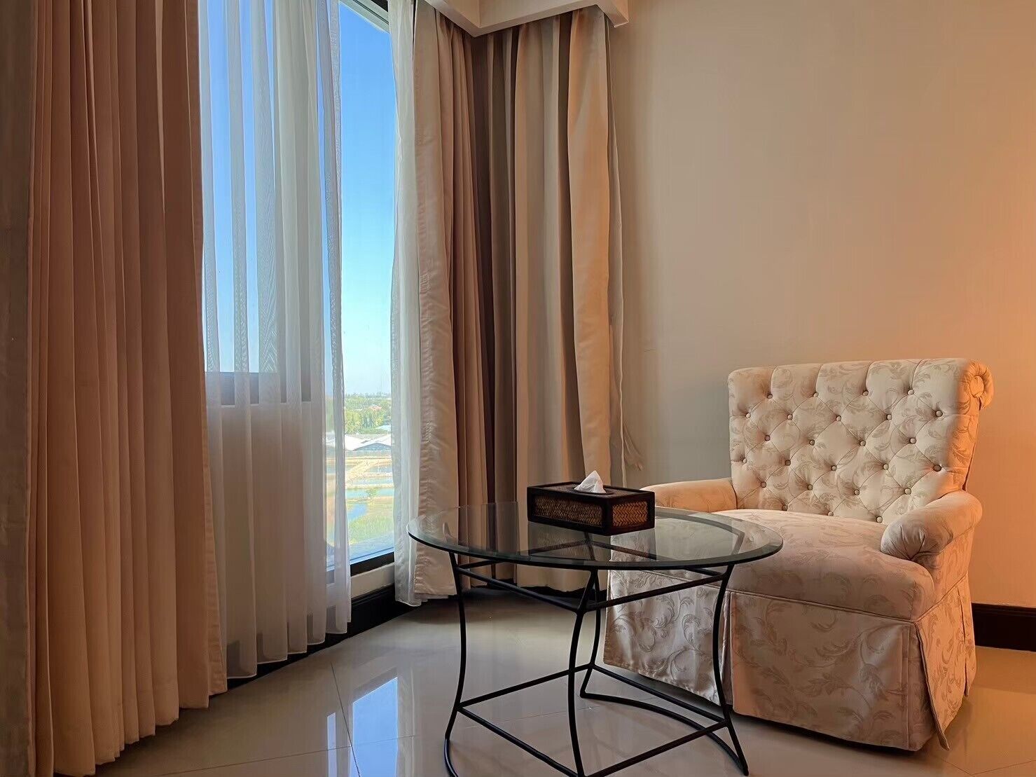 A room with a glass table, a tufted armchair, and a view through sheer curtains.