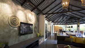 Un interior moderno con un televisor montado en una pared texturizada, una mesa de centro de madera con fruta, y un comedor con pared de ladrillo y luz natural.