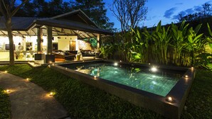 Zona junto a la piscina con un espacio de descanso cubierto, una piscina de piedra y abundante vegetación.
