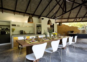 Uma sala de jantar espaçosa com uma longa mesa de madeira posta para seis pessoas, rodeada por cadeiras brancas. Uma parede de tijolos e uma cozinha moderna com forno embutido e pia são visíveis ao fundo.