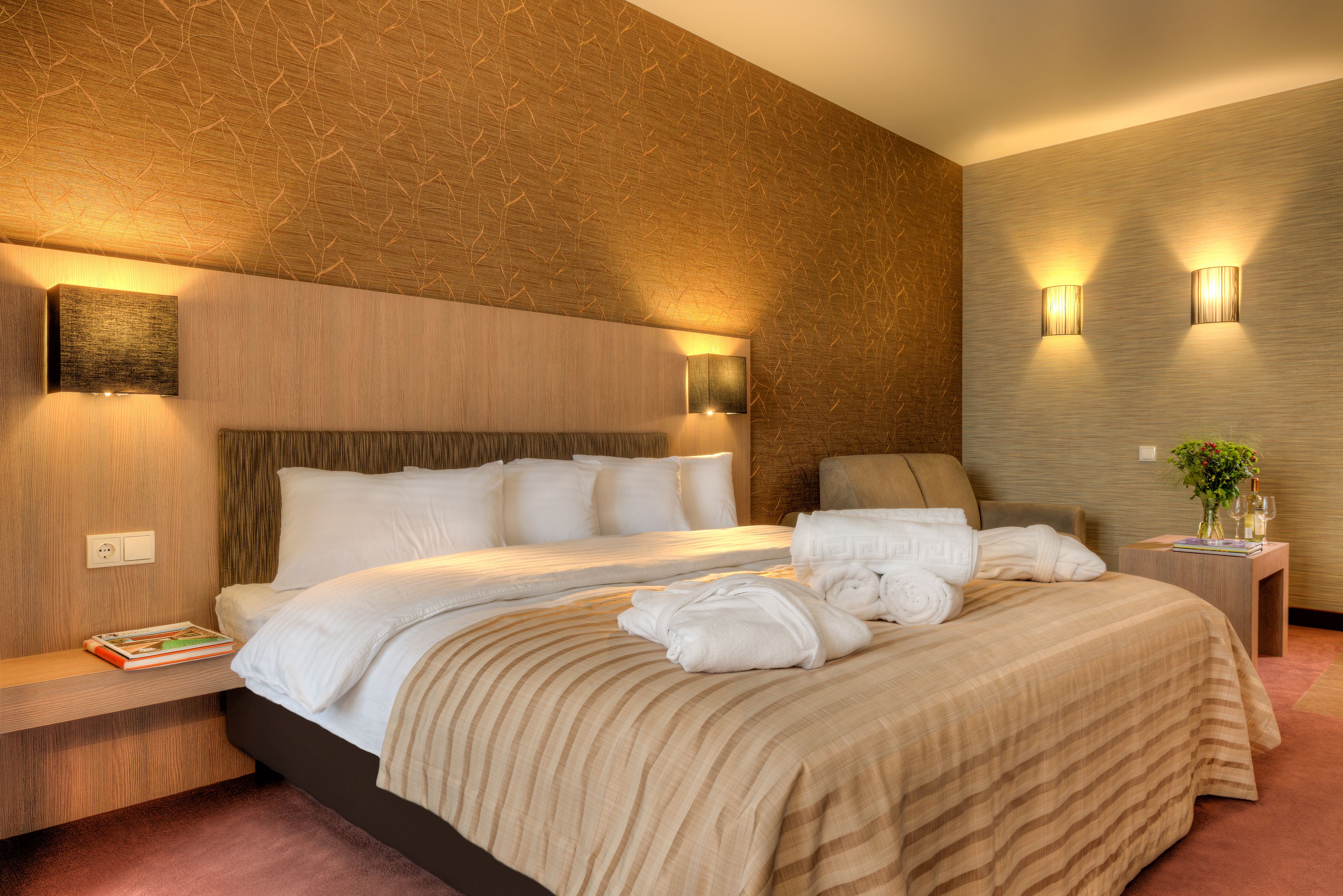 A modern hotel room with a large bed, a bedside table with a book, and two wall-mounted lamps.