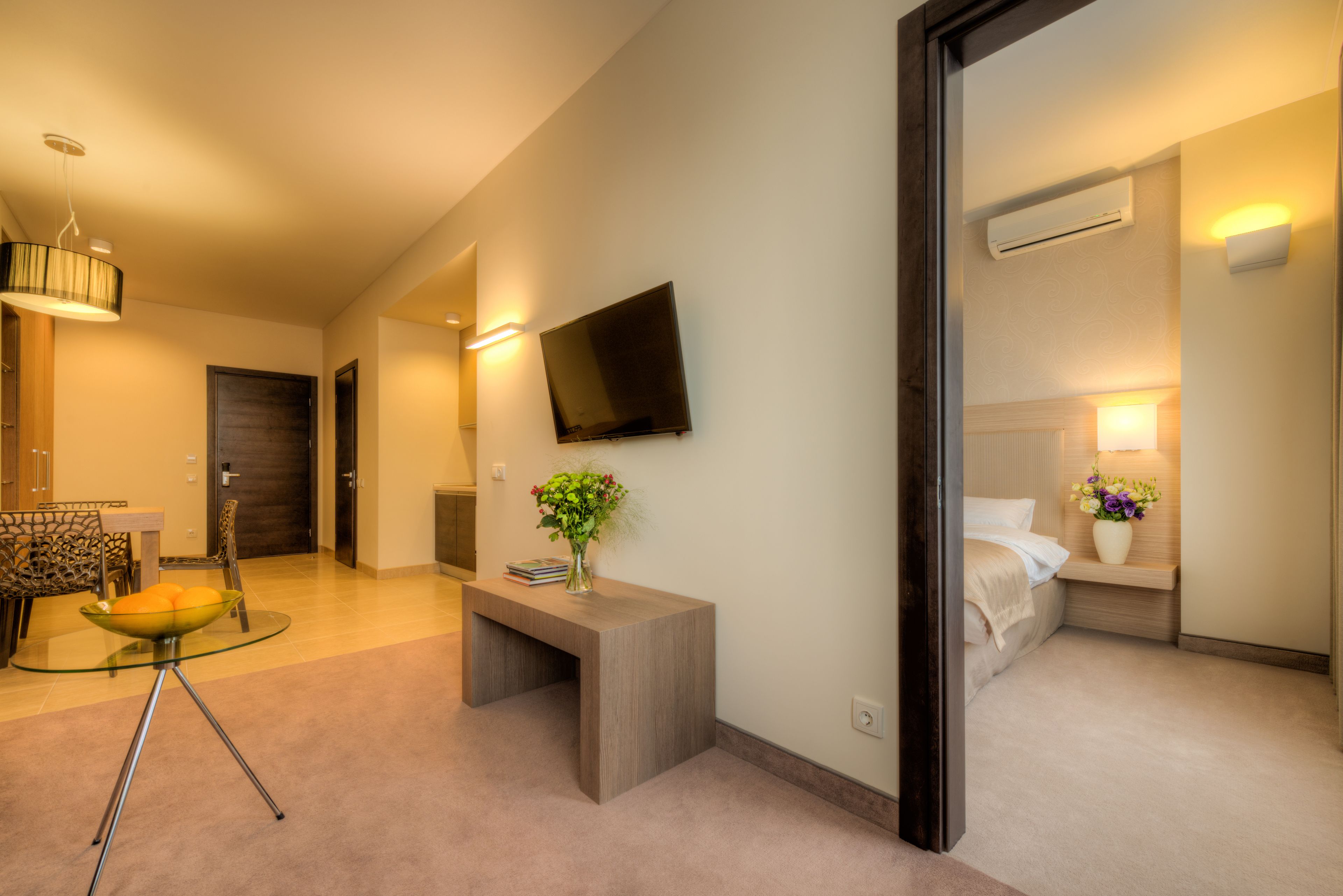 Ett modernt hotellrum med en platt-TV, ett sängbord med en vas med blommor, en säng med sänggavel och en liten sittgrupp med ett glasbord och stolar.