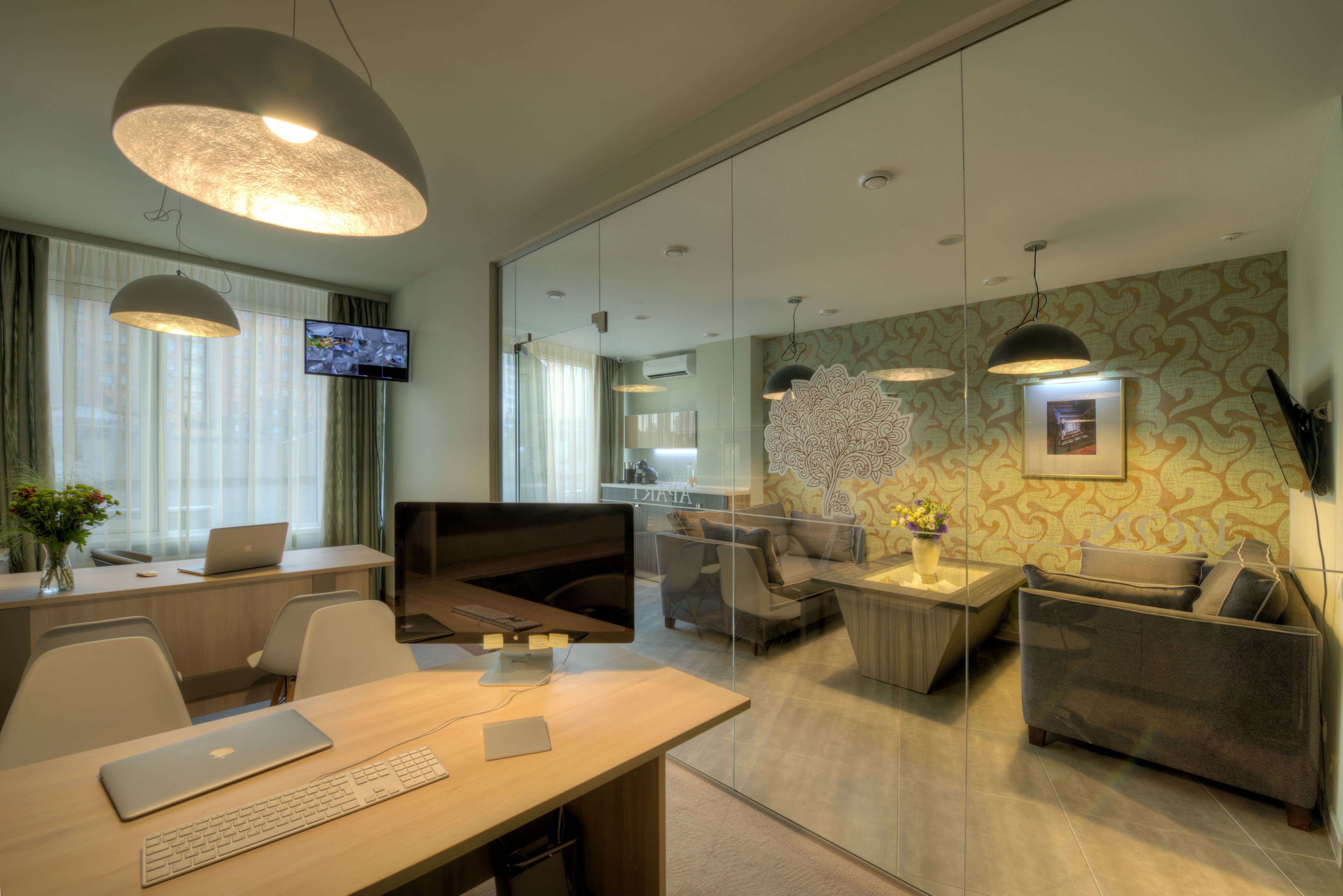 A modern office space with a desk, laptop, and a television. There is a sofa and a coffee table with a vase of flowers.