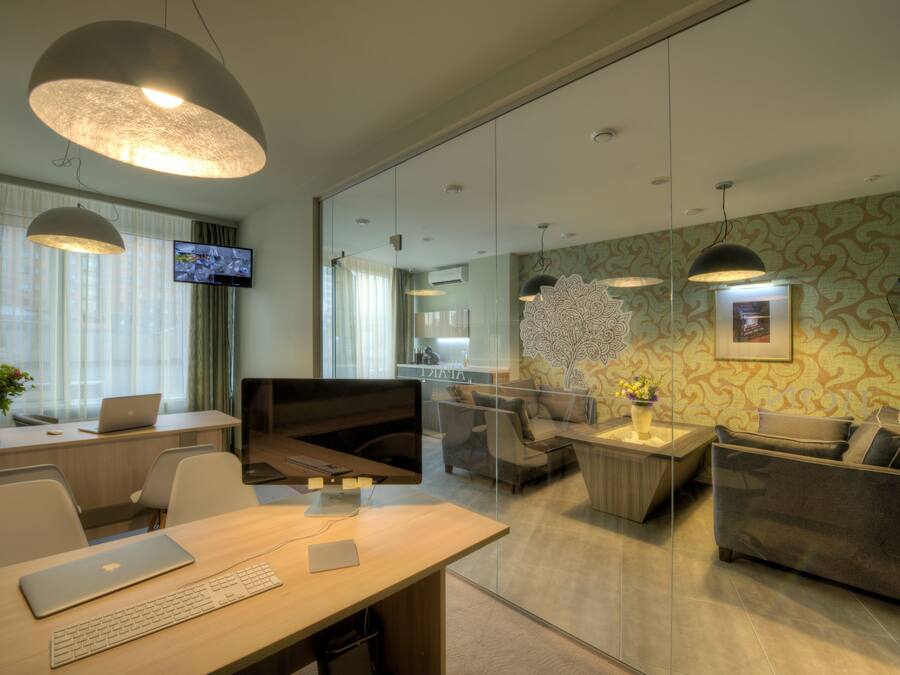 A modern office space with a desk, laptop, and a television. There is a sofa and a coffee table with a vase of flowers.