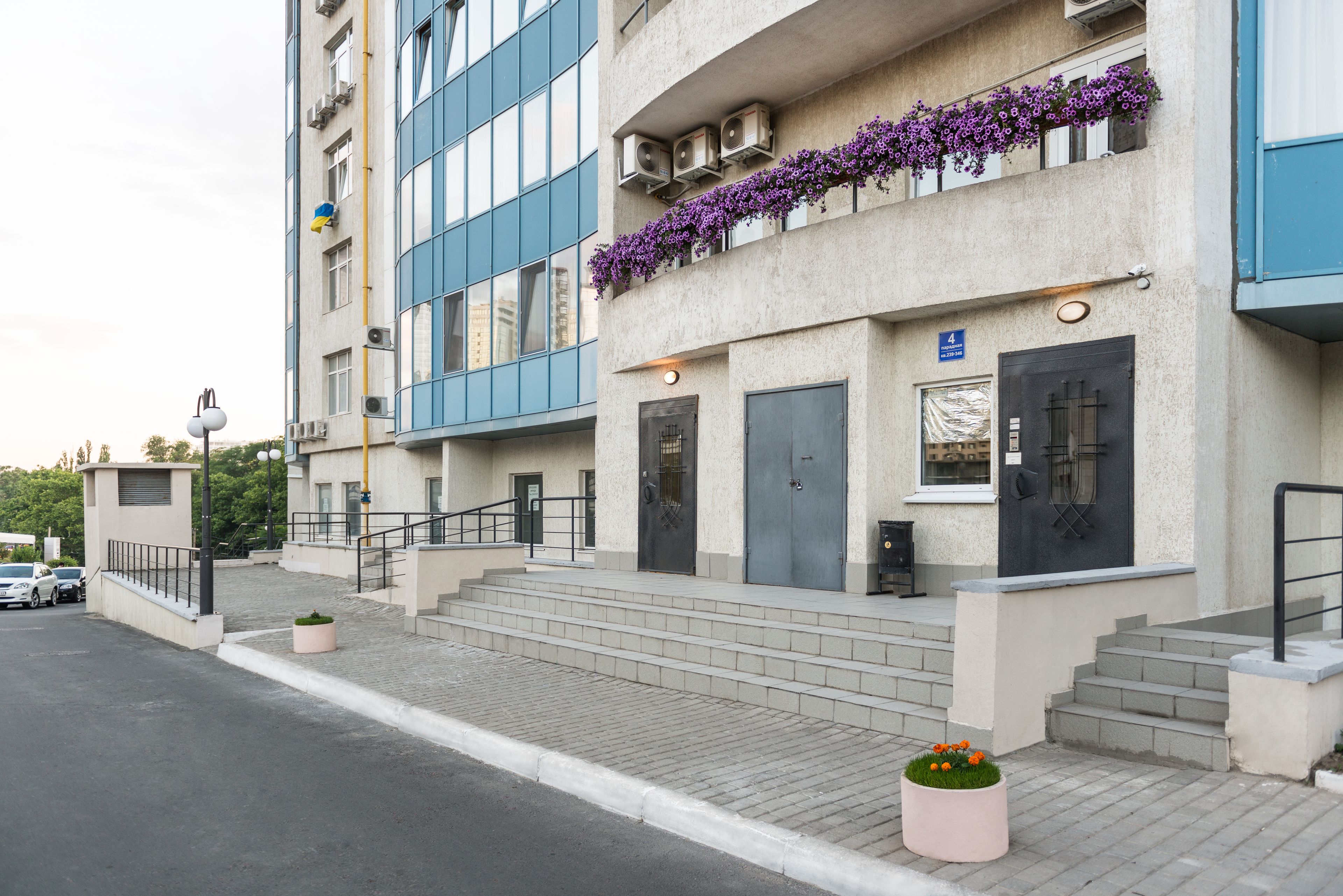 A multi-story building with a modern facade, featuring a set of stairs leading to the entrance, a small garden area with flowers, and a parking lot visible in the background.